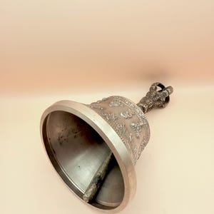 May include: A silver bell with a handle and intricate carvings. The bell is decorated with a repeating pattern of symbols and figures.
