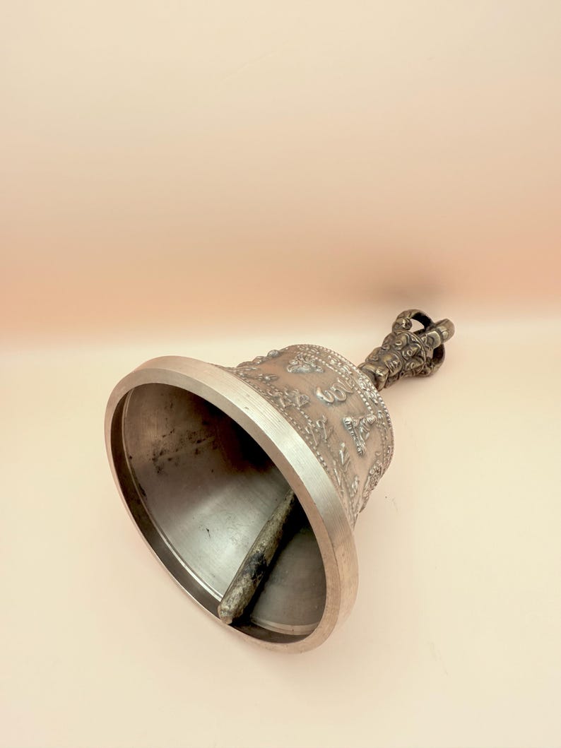 May include: A silver bell with a handle and intricate carvings. The bell is decorated with a repeating pattern of symbols and figures.
