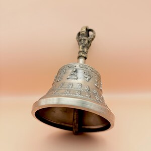 May include: A silver bell with a detailed handle and intricate engravings. The bell has a rounded shape and a wide opening. The handle is ornate and features a small figure at the top.