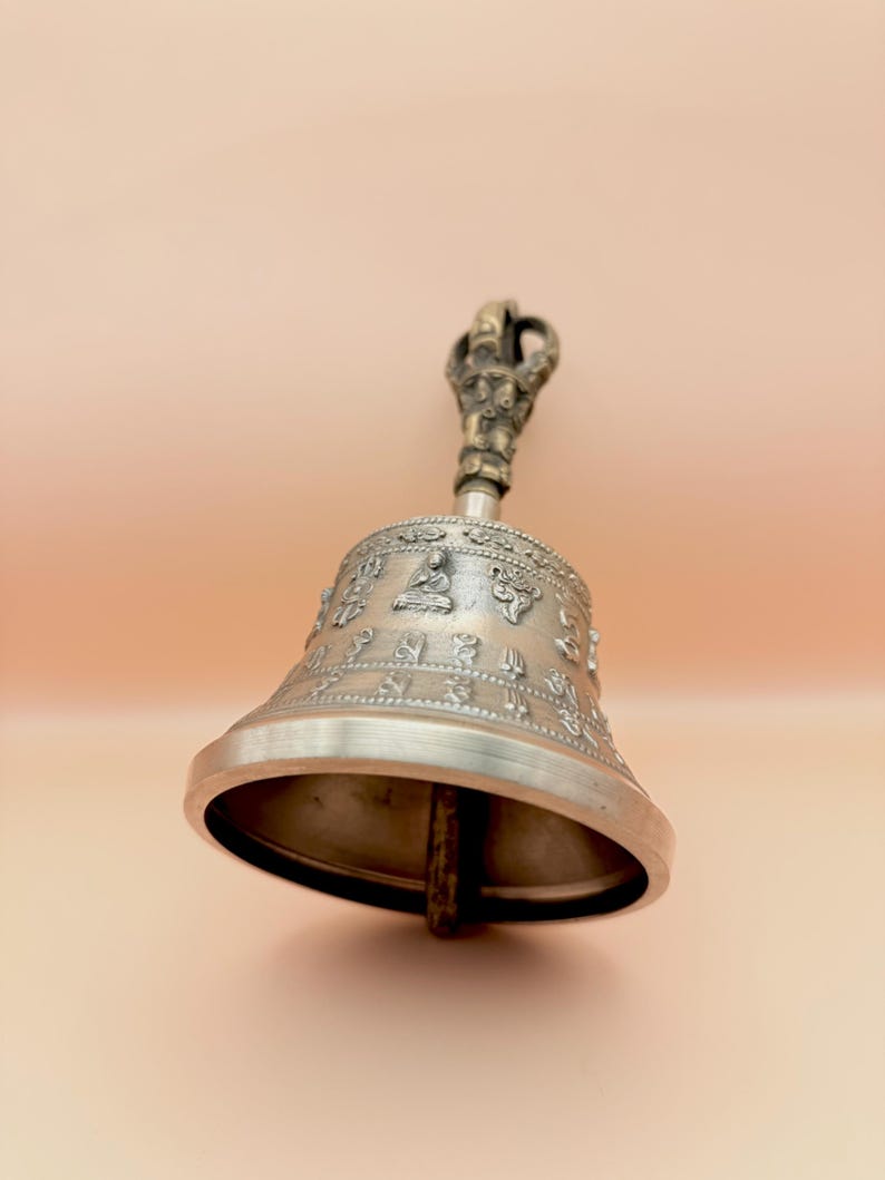 May include: A silver bell with a detailed handle and intricate engravings. The bell has a rounded shape and a wide opening. The handle is ornate and features a small figure at the top.