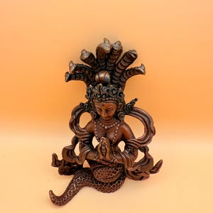 May include: A brown, ornate statue of a deity with a snake-like head and multiple snake heads as a crown. The deity is sitting with hands clasped in prayer and a snake tail.