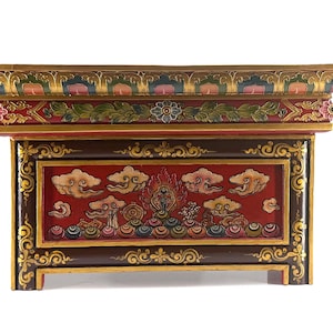 May include: A rectangular wooden altar table featuring a deep red and dark brown finish with intricate gold painted scrollwork. The front panel displays a carved relief of stylized clouds, a central flame motif, and colorful circular patterns.