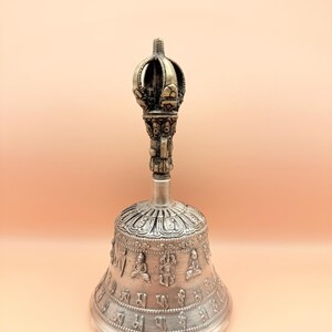May include: A silver bell with a decorative handle and intricate carvings. The bell is adorned with Sanskrit characters and small Buddha figures.