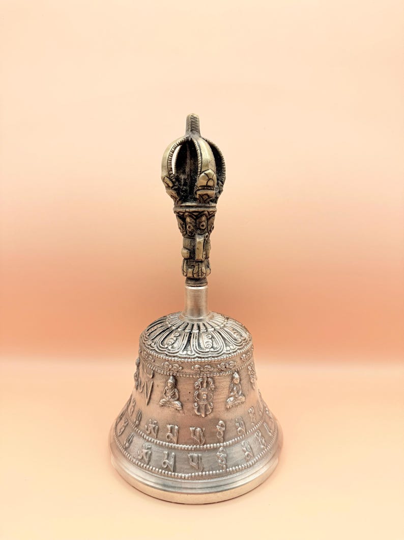 May include: A silver bell with a decorative handle and intricate carvings. The bell is adorned with Sanskrit characters and small Buddha figures.