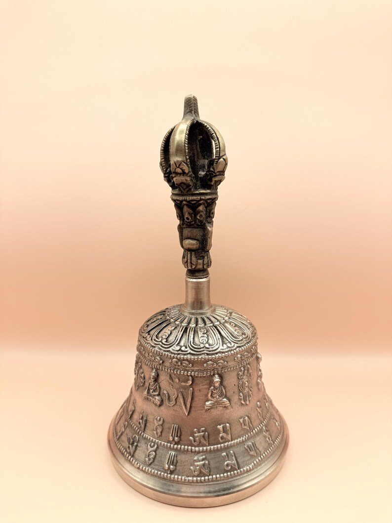 May include: A silver bell with intricate carvings and a crown-shaped handle. The bell is decorated with Buddhist symbols and figures.