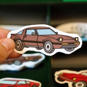 May include: A brown car sticker with a white border. The sticker features a cartoon illustration of a classic sports car with blue tinted windows and yellow headlights. The car has black stripes and gray wheels.