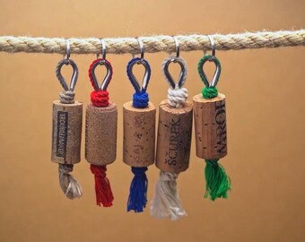May include: Five wine cork keychains with different colored strings hanging from a rope. The corks are labeled "TROUBLEMAKER", "SCUREK", "MAROF", and two unreadable labels.