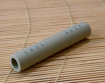 May include: A cylindrical, light gray object with a series of small, evenly spaced, silver-colored embellishments. The object has a hollow opening at one end. The background is a light brown bamboo mat.