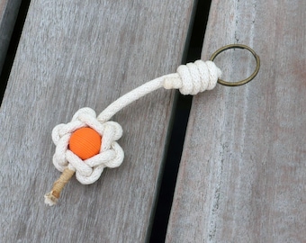 May include: A white rope keychain with a knotted flower design and an orange bead in the center. The keychain has a brass key ring.