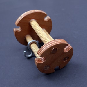 Puede incluir: Un enhebrador de lana de madera con dos clavijas y un gancho de metal. El enhebrador está hecho de madera contrachapada y tiene un acabado de madera natural.