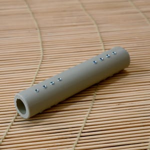 May include: A cylindrical, light gray object with a series of small, evenly spaced, silver-colored embellishments. The object has a hollow interior and is resting on a bamboo mat. The overall design is simple and modern.