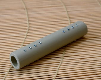 May include: A cylindrical, light gray object with a series of small, evenly spaced, silver-colored embellishments. The object has a hollow interior and is resting on a bamboo mat. The overall design is simple and modern.