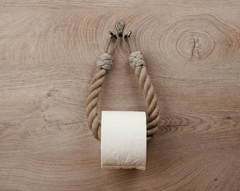 May include: A rope toilet paper holder with a roll of white toilet paper. The holder is made of natural rope and has a metal hook at the top.