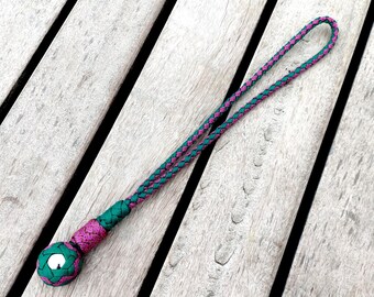 May include: A handcrafted lanyard featuring a braided cord in teal and purple. The lanyard has a decorative ball at the base with a white accent. The cord is looped at the top for easy wear. The lanyard is displayed on a wooden surface.