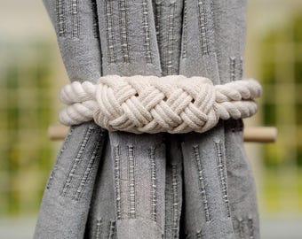 May include: A close-up of a white rope curtain tieback, tied around two gray curtains. The rope is knotted in a decorative pattern.