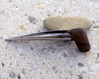 May include: A silver metal tool with a wooden handle. The tool has a pointed tip and a curved handle. The tool is lying on a gray concrete surface.