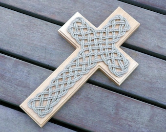 May include: A wooden cross with a rope design. The rope is woven into a pattern that resembles a Celtic knot. The cross is light brown and the rope is a light gray color.