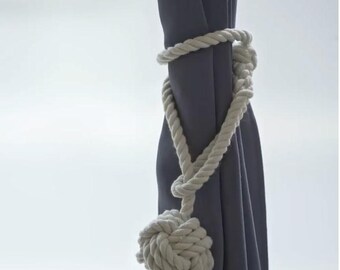 May include: A white cotton rope curtain tieback with a large knotted ball at the end. The tieback is wrapped around a gray curtain.