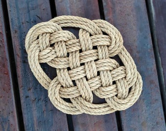 May include: A hand-woven, oval-shaped rope mat. The mat is made of thick, natural-colored rope, intricately woven in a basket-weave pattern. The mat rests on a dark wooden surface, creating a contrast in textures and tones. This nautical-themed mat is suitable for indoor or outdoor use.