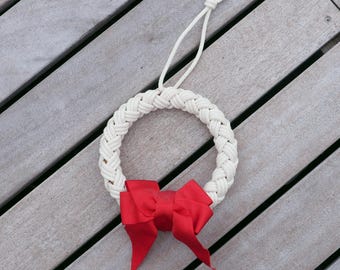May include: A white rope wreath with a red bow. The wreath is hanging on a wooden surface.