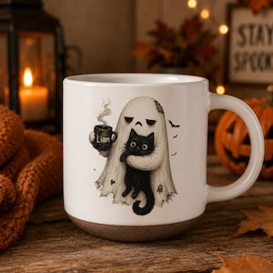 May include: A white ceramic mug featuring a printed illustration of a ghost draped in a sheet, holding a small black mug labeled Liam and embracing a fluffy black cat. The design includes small bats and hearts, with a brown base on the mug.