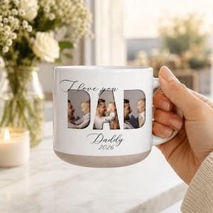 Könnte beinhalten: Weißer Keramikbecher mit hellbraunem Boden. Das Wort "DAD" ist ausgeschnitten und zeigt vier Fotos von Vater und Kind. Der Becher trägt den Text "I love you" darüber und "Daddy 2026" darunter. Eine Hand hält den Becher.