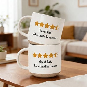 Könnte beinhalten: Zwei weiße Keramikbecher, die auf einem Holztisch gestapelt sind. Jeder Becher hat einen gesprenkelten Boden, einen gebogenen Henkel und den Text "Great Dad. Jokes could be funnier." über einer Reihe goldener Sterne.