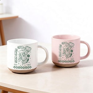 Könnte beinhalten: Zwei Keramikbecher, einer weiß und einer rosa, mit braunem Boden und Henkel. Jeder Becher zeigt ein grünes Cartoon-Design eines Bierkrugs mit dem Text "COOL DADS CLUB ESTD 2026". Die Becher stehen auf einer weißen Oberfläche.