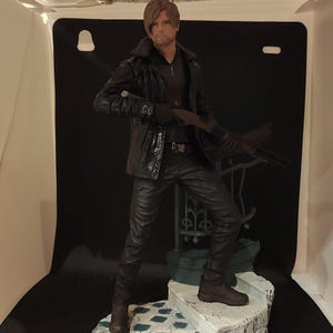 May include: A detailed figurine of a man in a black leather jacket and pants, holding a rifle. The figure stands on a detailed, weathered base with a distressed, painted finish. The figure is a collectible item.