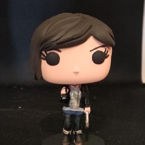 May include: A Funko Pop vinyl figure of a character with short brown hair, wearing a black jacket, gray shirt, blue jeans, and black boots. The figure is holding a silver object in one hand and stands on a black circular base.