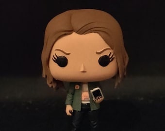 May include: A vinyl collectible figure with brown hair, large black eyes, and a small nose. The figure wears an olive green jacket over a white top, black pants, and dark boots, while holding a small black rectangular device in one hand.
