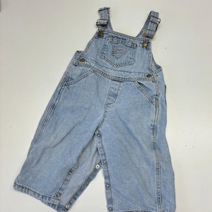 Könnte beinhalten: Hellblaue Jeans-Latzhosen mit verstellbaren Trägern und einem Guess Originals Logo-Patch auf der Vorderseite. Die Latzhosen haben einen Druckknopfverschluss unten.