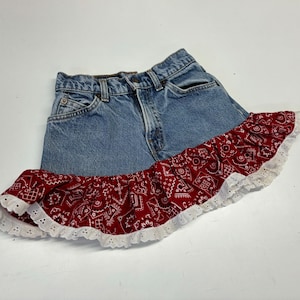 May include: A denim skirt with a red bandana-style frill. The skirt is made of blue denim and features a red fabric with a white paisley pattern. The frill is trimmed with white eyelet lace.