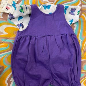 May include: A purple overall romper with a white short-sleeved shirt featuring a butterfly print. The shirt has a white background with blue and purple butterflies.
