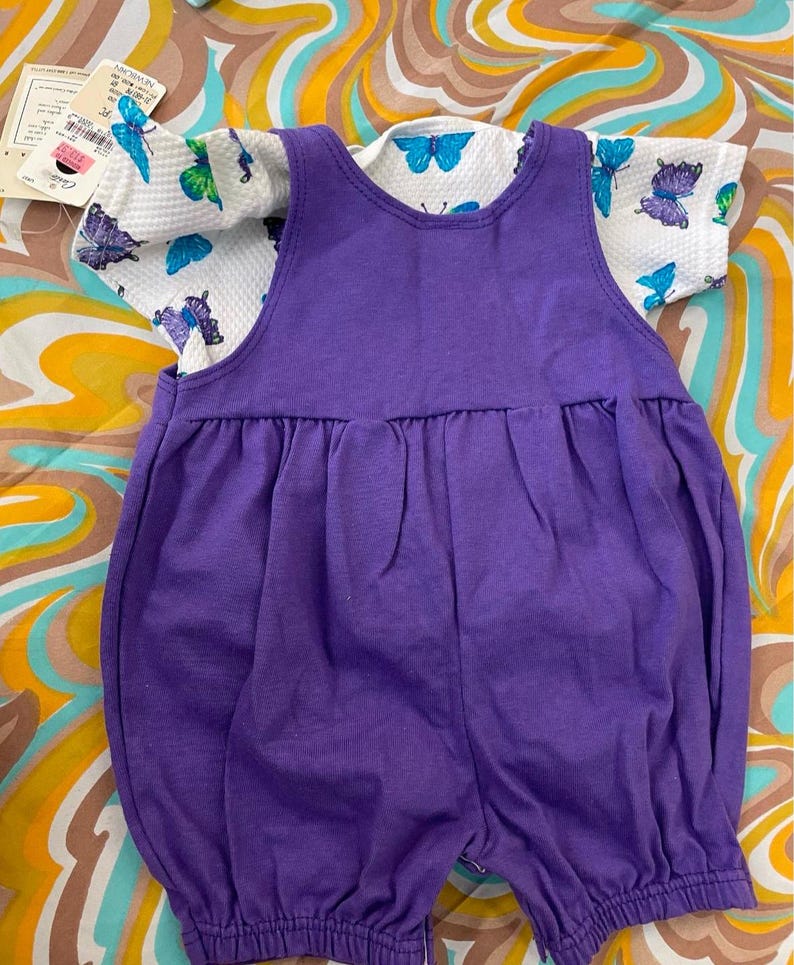 May include: A purple overall romper with a white short-sleeved shirt featuring a butterfly print. The shirt has a white background with blue and purple butterflies.
