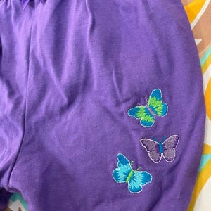 May include: Purple shorts with embroidered butterflies in shades of green, blue, and purple.