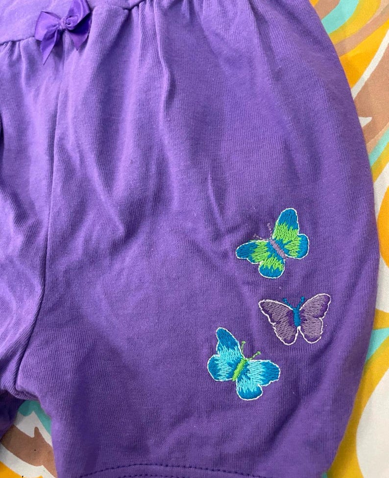 May include: Purple shorts with embroidered butterflies in shades of green, blue, and purple.