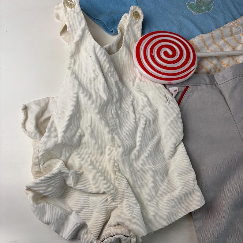 K&ouml;nnte beinhalten: Eine Auswahl an Kinderkleidung, darunter ein cremefarbener Cord-Strampler mit Knopfverschl&uuml;ssen, ein hellblaues Oberteil und graue Shorts. Ein rot-wei&szlig;es Wirbellutscher liegt auf der Kleidung.
