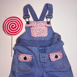 May include: Blue denim overalls with floral print accents on the pockets and bib. A large red and white swirled lollipop is positioned to the left of the overalls. The overalls have adjustable straps and button closures.