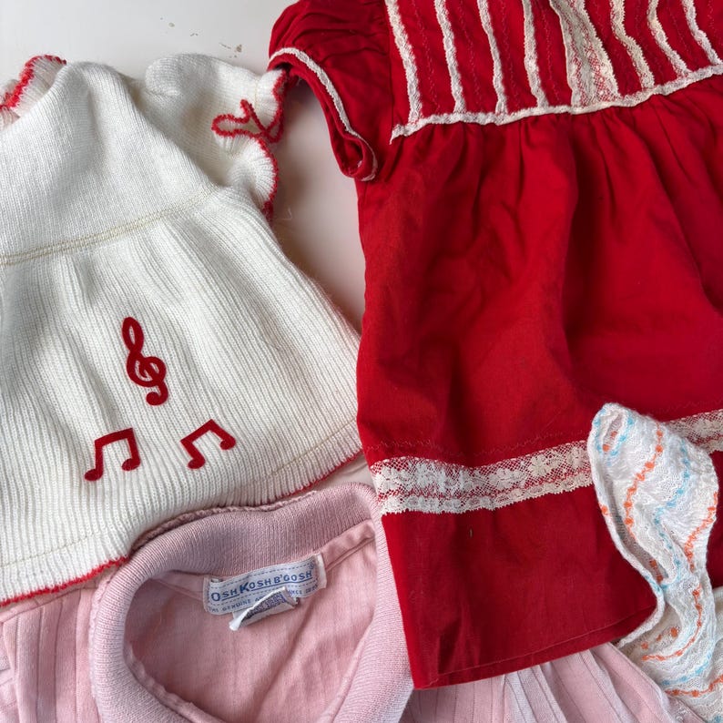 K&ouml;nnte beinhalten: Eine Sammlung von Vintage-Kinderkleidung. Enth&auml;lt ein wei&szlig;es Strickoberteil mit roten Noten, ein rotes Kleid mit wei&szlig;er Spitzenborte und ein rosa Hemd mit Kragen. Das Etikett OshKosh B'gosh ist auf dem rosa Hemd zu sehen.