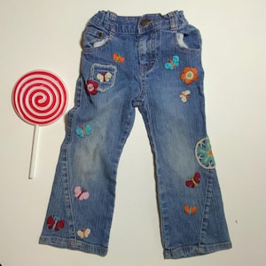 Może przedstawiać: Niebieskie dżinsy dziecięce ozdobione kolorowymi haftowanymi motylami i kwiatami. Dżinsy mają klasyczny krój z pięcioma kieszeniami i zapięciem na guziki. Po lewej stronie znajduje się czerwono-biały lizak.