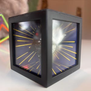 Puede incluir: Una caja de presentación de forma cúbica negra con un panel frontal transparente. La caja presenta una imagen 3D de Darth Vader y Yoda, con un patrón de estrella amarilla sobre un fondo azul oscuro.