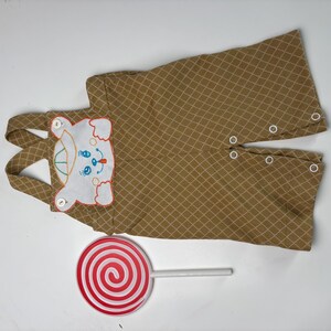 K&ouml;nnte beinhalten: Ein beigefarbener Baby-Strampler mit einem wei&szlig;en, gestickten B&auml;rengesicht auf der Brust. Der Strampler hat ein Rautenmuster und wei&szlig;e Kn&ouml;pfe. Ein gro&szlig;er rot-wei&szlig; gestreifter Lutscher liegt daneben.