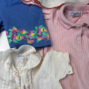 K&ouml;nnte beinhalten: Eine Auswahl an Kinderkleidung, darunter ein blaues Kurzarmshirt mit floraler Stickerei, ein rosa geripptes Poloshirt und ein wei&szlig;es Hemd mit Kn&ouml;pfen. Die Artikel sind in verschiedenen Blau-, Rosa- und Wei&szlig;t&ouml;nen gehalten.