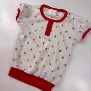 Puede incluir: Una camiseta blanca de estilo vintage con un estampado floral en rojo, azul, amarillo y morado. La camiseta tiene un cuello rojo, mangas cortas y una banda roja en la cintura. Dos botones rojos están en la parte delantera.