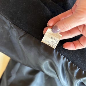 K&ouml;nnte beinhalten: Nahaufnahme des Innenraums eines schwarzen Mantels, der ein Etikett mit dem Text "GIRLS SIZE S" und der Losnummer "MX048-5" zeigt. Das Futter des Mantels ist ein gl&auml;nzendes, dunkles Material.