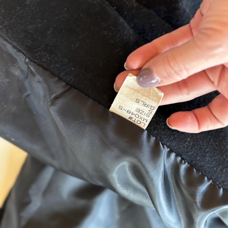 K&ouml;nnte beinhalten: Nahaufnahme des Innenraums eines schwarzen Mantels, der ein Etikett mit dem Text "GIRLS SIZE S" und der Losnummer "MX048-5" zeigt. Das Futter des Mantels ist ein gl&auml;nzendes, dunkles Material.