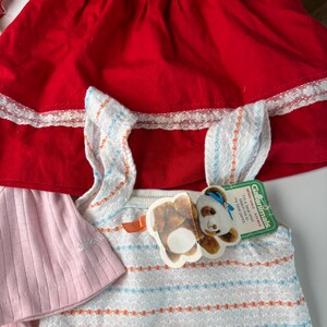 K&ouml;nnte beinhalten: Ein rotes Kleid mit wei&szlig;er Spitzenborte, ein rosa Oberteil und ein wei&szlig;-blau gestreiftes Hemd mit einem B&auml;renanh&auml;nger. Auf dem Anh&auml;nger steht "Garanimals" und "I'm a bear shirt. Match me to my bear pants."