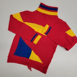 May include: Red turtleneck sweater with yellow and blue geometric patterns. The sweater has a contrasting blue, yellow, and red stripe at the neck.