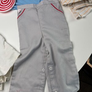 K&ouml;nnte beinhalten: Graue Babyhose mit roter Paspel an den Taschen. Die Hose hat Druckkn&ouml;pfe am unteren Bein. Die Hose ist aus weichem Material und wahrscheinlich Vintage.